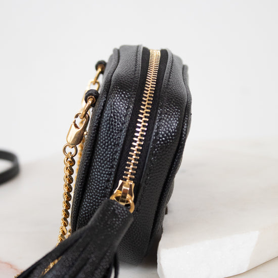 A close-up side view of a black textured leather crossbody bag with gold-tone hardware, a tassel detail, and a gold chain strap, resting on a white surface.