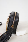 A close-up side view of a black textured leather crossbody bag with gold-tone hardware, a tassel detail, and a gold chain strap, resting on a white surface.