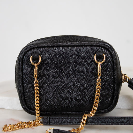 A black textured leather camera bag with gold-tone chain strap details, resting on a light marble surface against a white background.