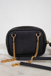 A black textured leather camera bag with gold-tone chain strap details, resting on a light marble surface against a white background.