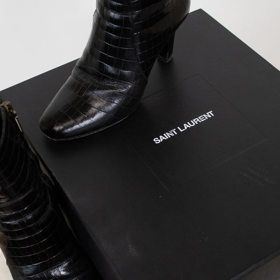 A pair of black Saint Laurent crocodile-embossed ankle boots displayed next to their black Saint Laurent box on a light marble surface.