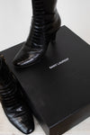 A pair of black Saint Laurent crocodile-embossed ankle boots displayed next to their black Saint Laurent box on a light marble surface.
