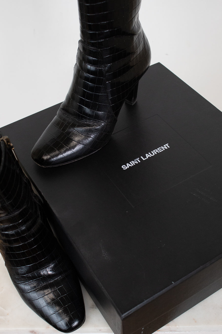 A pair of black Saint Laurent crocodile-embossed ankle boots displayed next to their black Saint Laurent box on a light marble surface.