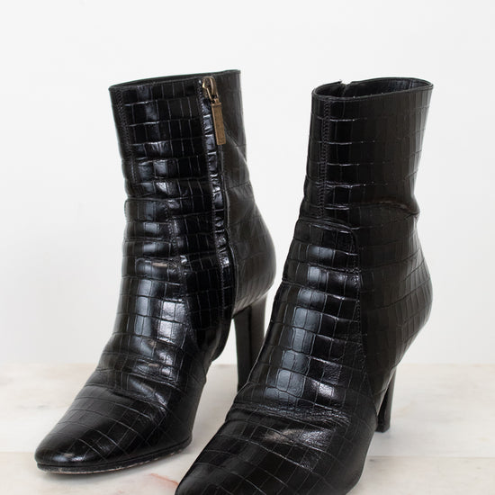 A pair of black crocodile-embossed leather ankle boots with pointed toes and kitten heels, displayed on a light marble surface against a white background.