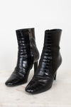 A pair of black crocodile-embossed leather ankle boots with pointed toes and kitten heels, displayed on a light marble surface against a white background.