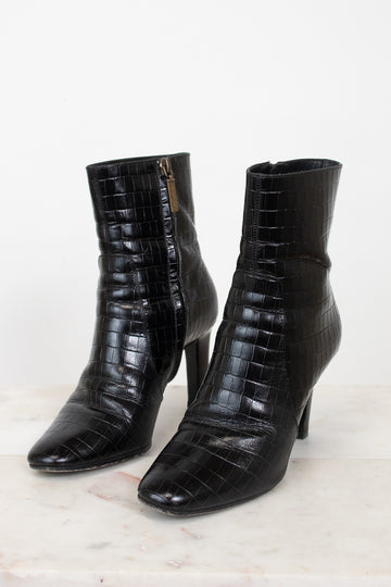 A pair of black crocodile-embossed leather ankle boots with pointed toes and kitten heels, displayed on a light marble surface against a white background.