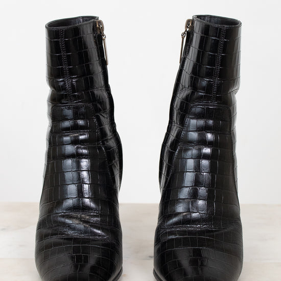 A pair of black crocodile-embossed leather ankle boots with side zippers, displayed on a light marble surface against a white background.
