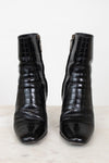 A pair of black crocodile-embossed leather ankle boots with side zippers, displayed on a light marble surface against a white background.