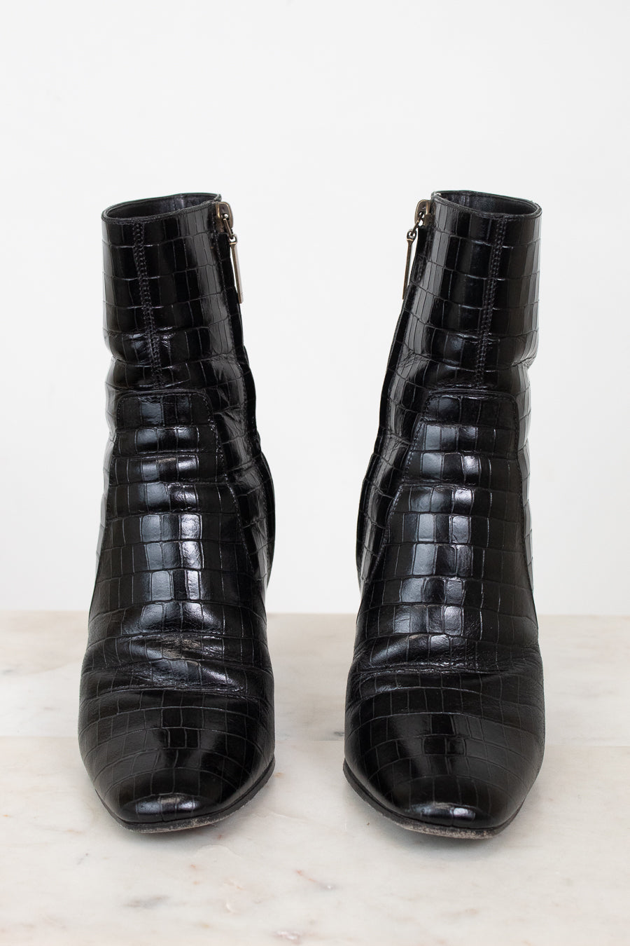 A pair of black crocodile-embossed leather ankle boots with side zippers, displayed on a light marble surface against a white background.