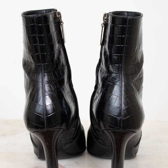 A pair of black high-heeled ankle boots with an embossed crocodile pattern, shown from the back on a light marble surface against a white background.