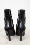 A pair of black high-heeled ankle boots with an embossed crocodile pattern, shown from the back on a light marble surface against a white background.