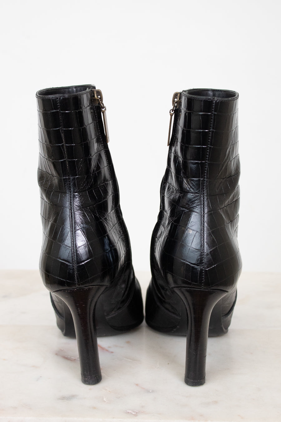 A pair of black high-heeled ankle boots with an embossed crocodile pattern, shown from the back on a light marble surface against a white background.
