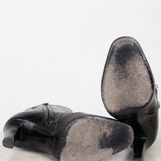 A pair of black leather high-heeled shoes shown from the bottom, revealing heavily worn bottom soles, set against a plain white background.