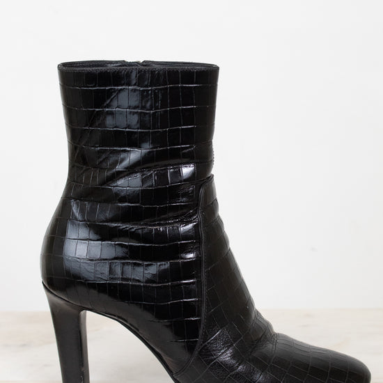 A single black ankle boot with a glossy crocodile-embossed texture and a high block heel, shown from a side profile against a white background.