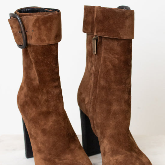 A pair of brown suede ankle boots with a chunky black block heel and a wide strap detail with a rhodium buckle on the back, shown from the rear on a light marble surface against a white background.
