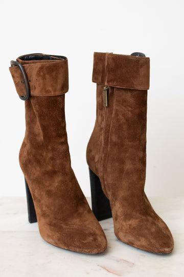A pair of brown suede ankle boots with a chunky black block heel and a wide strap detail with a rhodium buckle on the back, shown from the rear on a light marble surface against a white background.