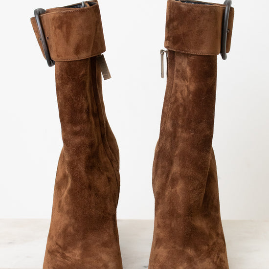 A pair of brown suede ankle boots with a chunky black block heel and a wide strap detail with a rhodium buckle on the back, shown from the rear on a light marble surface against a white background.