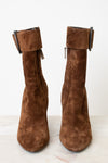 A pair of brown suede ankle boots with a chunky black block heel and a wide strap detail with a rhodium buckle on the back, shown from the rear on a light marble surface against a white background.