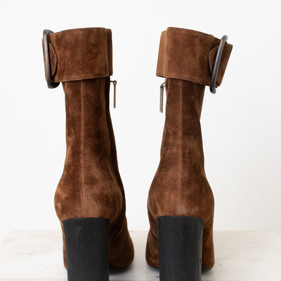 A pair of brown suede ankle boots with a chunky black block heel and a wide strap detail with a rhodim buckle on the back, shown from the rear on a light marble surface against a white background.