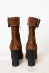 A pair of brown suede ankle boots with a chunky black block heel and a wide strap detail with a rhodim buckle on the back, shown from the rear on a light marble surface against a white background.