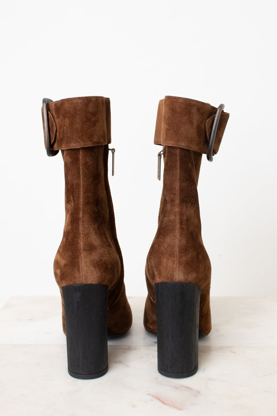 A pair of brown suede ankle boots with a chunky black block heel and a wide strap detail with a rhodim buckle on the back, shown from the rear on a light marble surface against a white background.