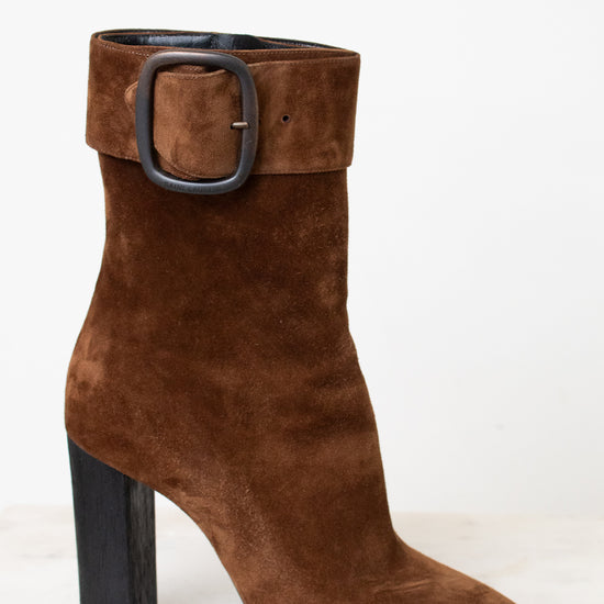 A brown suede ankle boot with a chunky black block heel and a wide strap detail with a silver buckle on the back, shown from the rear on a light marble surface against a white background.