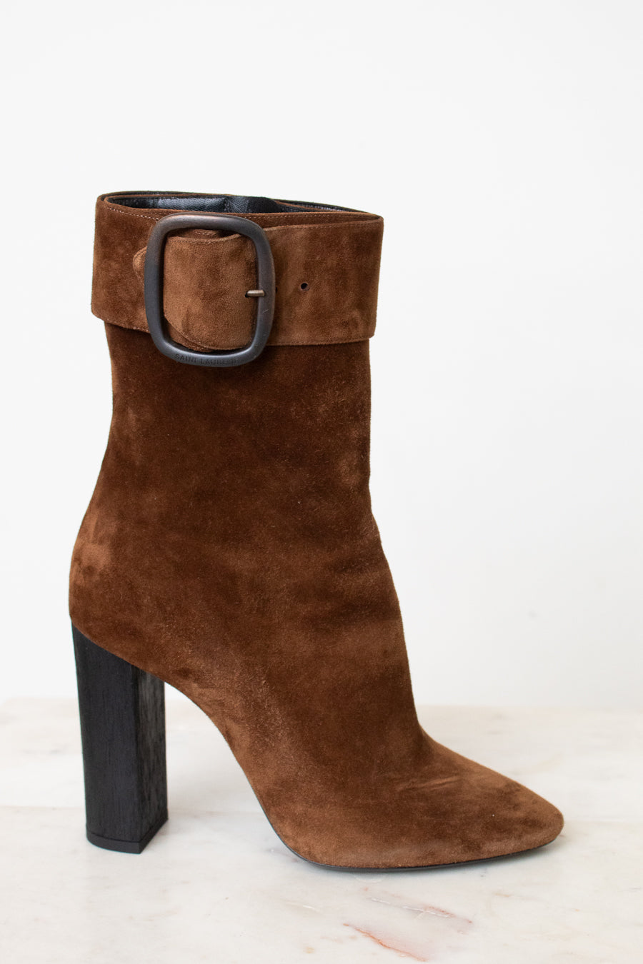 A brown suede ankle boot with a chunky black block heel and a wide strap detail with a silver buckle on the back, shown from the rear on a light marble surface against a white background.
