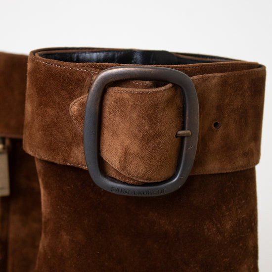 A pair of brown suede ankle boots with a chunky black block heel and a wide strap detail with a silver buckle on the back, shown from the rear on a light marble surface against a white background.