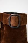 A pair of brown suede ankle boots with a chunky black block heel and a wide strap detail with a silver buckle on the back, shown from the rear on a light marble surface against a white background.