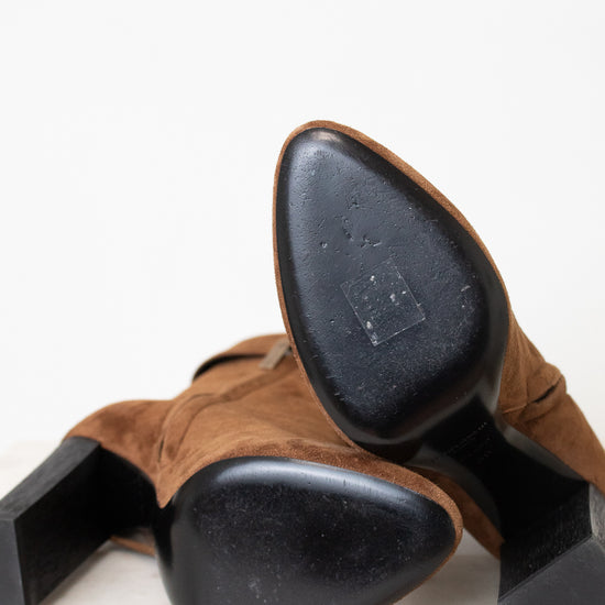 A pair of brown suede ankle boots with a chunky black block heel and black sole sitting on a light marble surface against a white background.