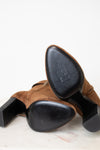 A pair of brown suede ankle boots with a chunky black block heel and black sole sitting on a light marble surface against a white background.