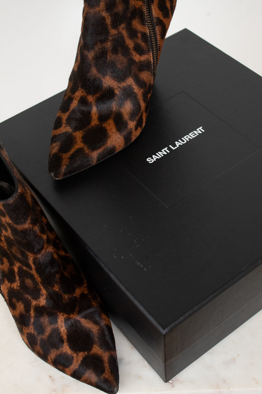 Saint Laurent leopard-print pointed-toe ankle boots displayed on a black Saint Laurent shoebox against a light marble background.