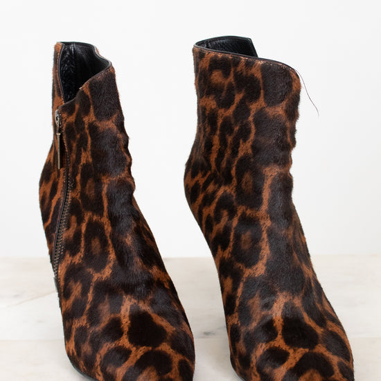 A pair of pointed-toe ankle boots with a brown and black leopard print pattern, shown on a light marble surface against a white background.