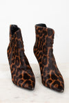 A pair of pointed-toe ankle boots with a brown and black leopard print pattern, shown on a light marble surface against a white background.