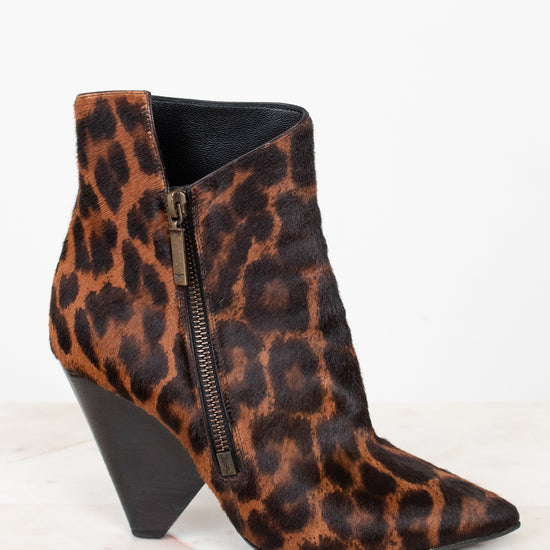 A pointed-toe ankle boot with a brown and black leopard print pattern, featuring a black conical heel and a side zipper, displayed on a light marble surface against a white background.
