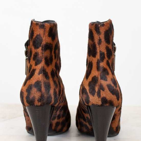 A pair of leopard print ankle boots with black conical heels, shown from the back on a light marble surface against a white background.