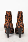 A pair of leopard print ankle boots with black conical heels, shown from the back on a light marble surface against a white background.