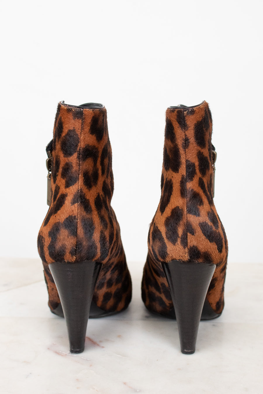 A pair of leopard print ankle boots with black conical heels, shown from the back on a light marble surface against a white background.
