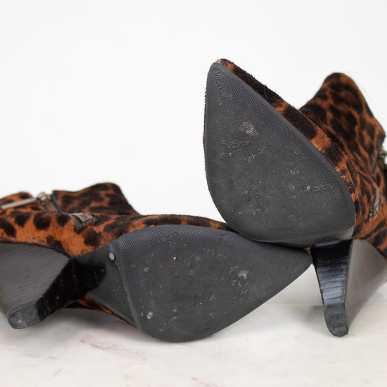 A pair of pointed-toe ankle boots with a brown and black leopard print, shown from the bottom on a white marble surface against a white background.