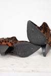 A pair of pointed-toe ankle boots with a brown and black leopard print, shown from the bottom on a white marble surface against a white background.
