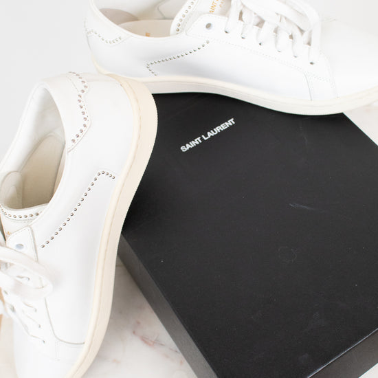 A pair of white leather Saint Laurent sneakers displayed next to their signature black box on a light marble surface.
