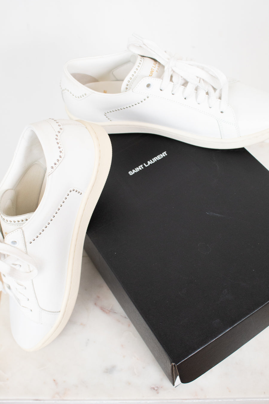 A pair of white leather Saint Laurent sneakers displayed next to their signature black box on a light marble surface.