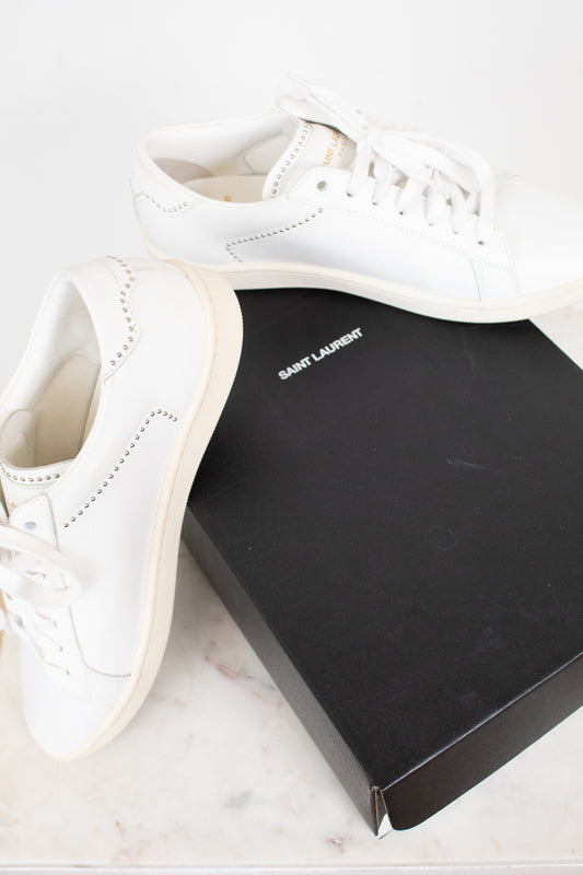 A pair of white leather Saint Laurent sneakers displayed next to their signature black box on a light marble surface.