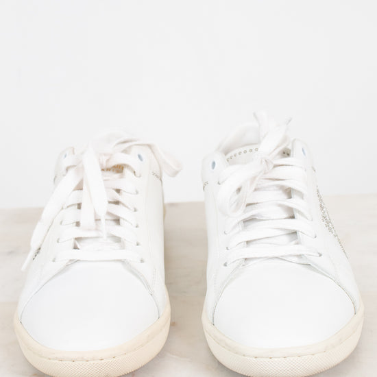 A pair of white leather low-top sneakers with white laces, resting on a light marble surface against a white background.