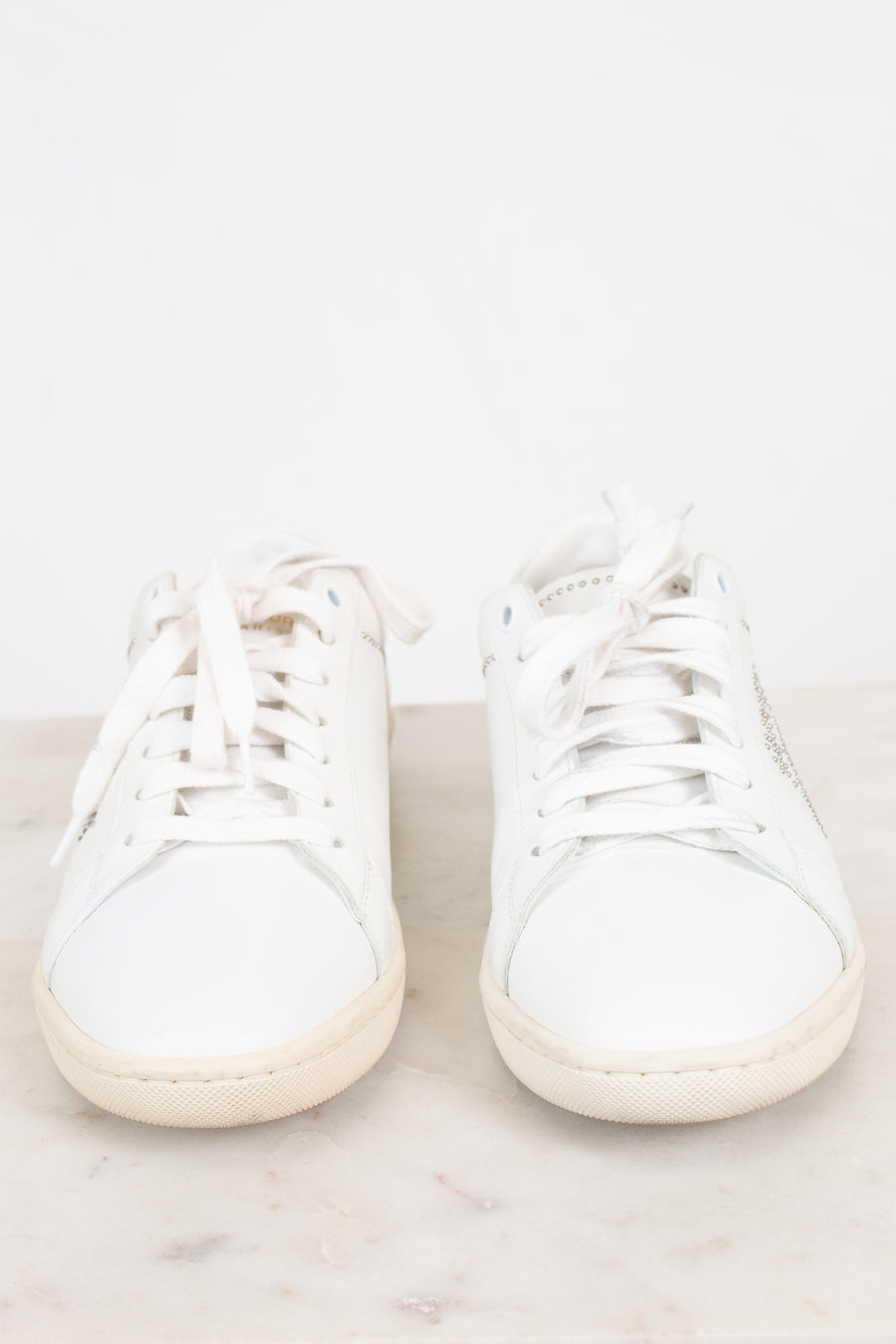 A pair of white leather low-top sneakers with white laces, resting on a light marble surface against a white background.