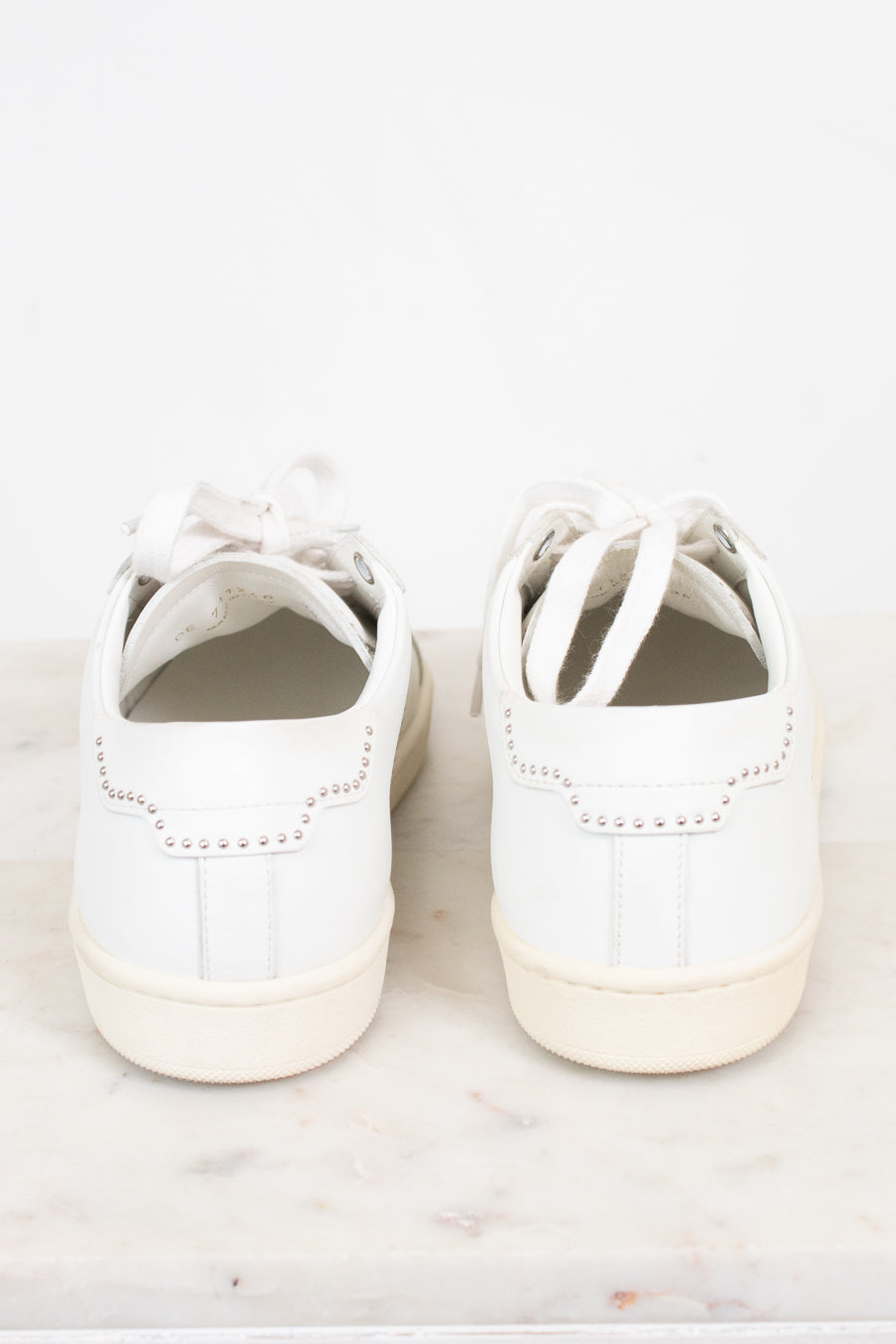 A pair of white leather low-top sneakers shown from the back, resting on a light marble surface against a white background.
