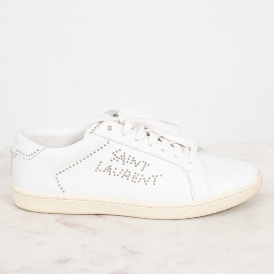A white leather Saint Laurent sneaker with silver rhinestone lettering on the side, displayed on a light marble surface against a white background.