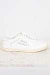 A white leather Saint Laurent sneaker with silver rhinestone lettering on the side, displayed on a light marble surface against a white background.