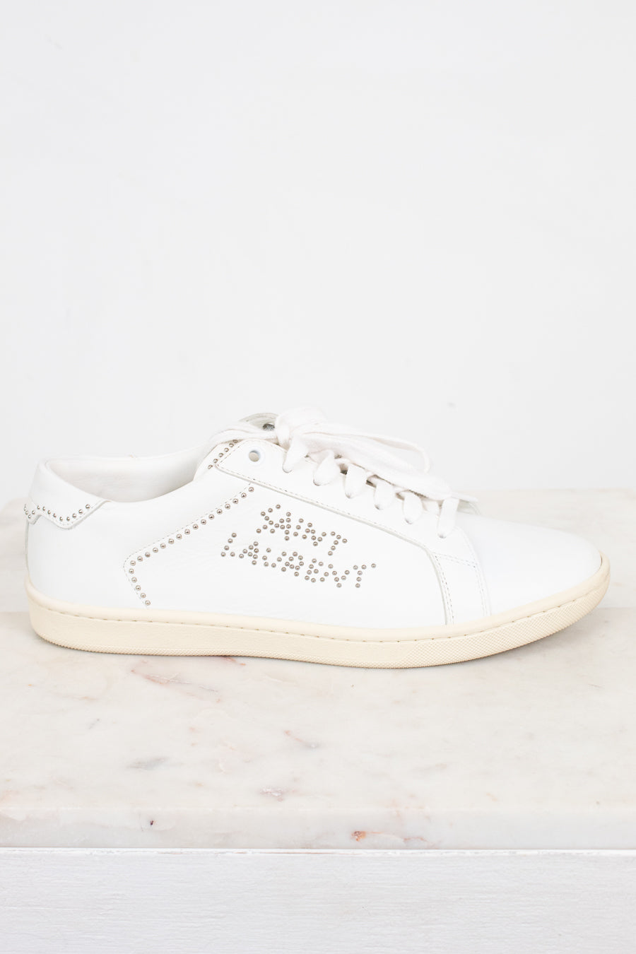 A white leather Saint Laurent sneaker with silver rhinestone lettering on the side, displayed on a light marble surface against a white background.