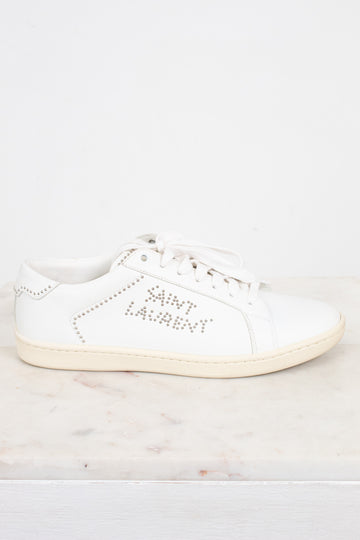 A white leather Saint Laurent sneaker with silver rhinestone lettering on the side, displayed on a light marble surface against a white background.